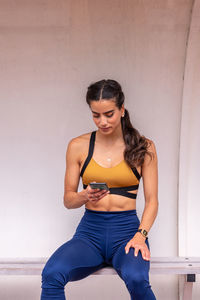 Young athletic hispanic sportswoman in activewear sitting on bench and using mobile app on smartphone while resting after training