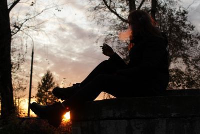 Side view of silhouette woman sitting on tree at sunset