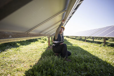 Female engineer repairing solar panels while kneeling on grass at power station