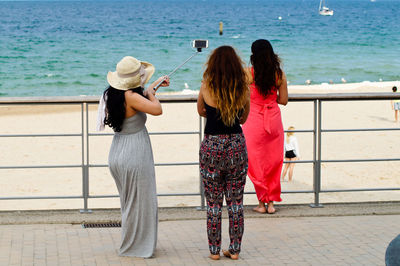 Rear view of friends taking selfie while standing by railing against sea