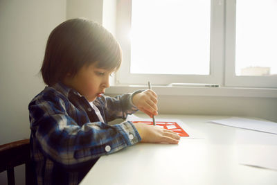 Boy child draws on paper with a ruler on a table sitting by the window