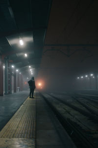 Back view of unrecognizable tourist with backpack waiting for train on platform under shiny lamps at dusk