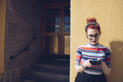 Young female hipster using mobile phone while standing against wall by entrance of building