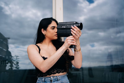 Young woman holding camera while standing against sky