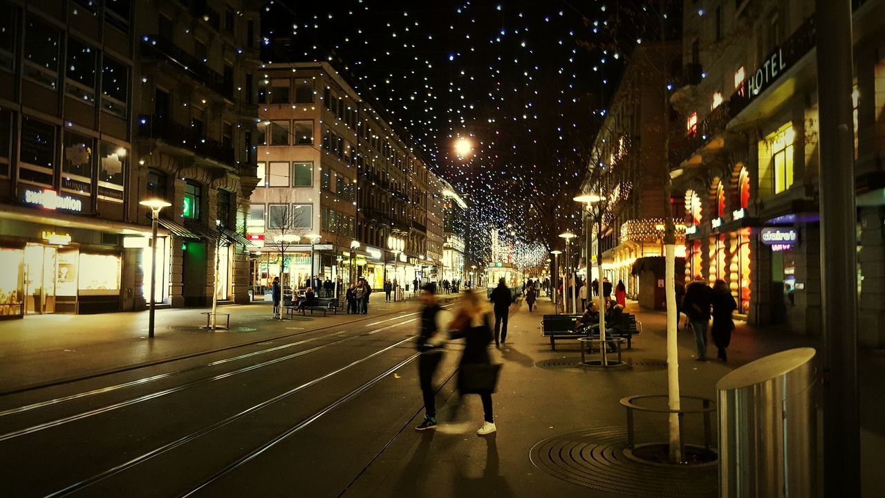 illuminated, night, building exterior, city, architecture, built structure, walking, street, large group of people, men, person, city life, lifestyles, lighting equipment, city street, leisure activity, street light, building, group of people