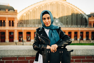 Portrait of young woman standing against wall in city