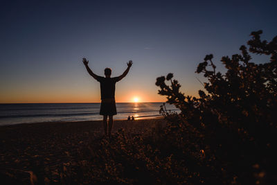 Rear view of man with arms raised standing at beach during sunset