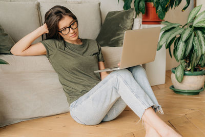 A girl in glasses watches a video in a computer while sitting on the floor.