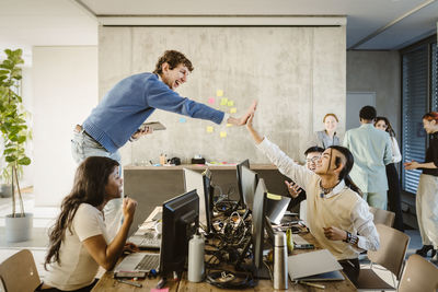 Cheerful entrepreneurs giving high-five while colleagues sitting at desk in creative office