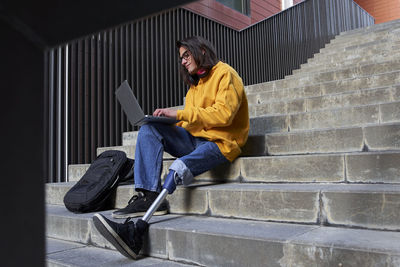 Disabled young man with leg prosthesis using laptop while sitting on steps