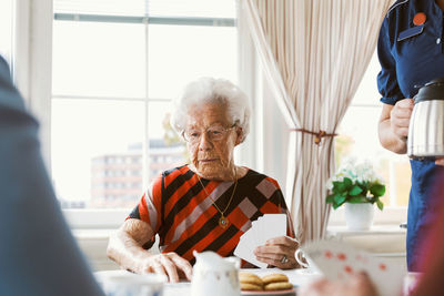 Senior woman playing cards with family while caretaker holding coffee pot at home
