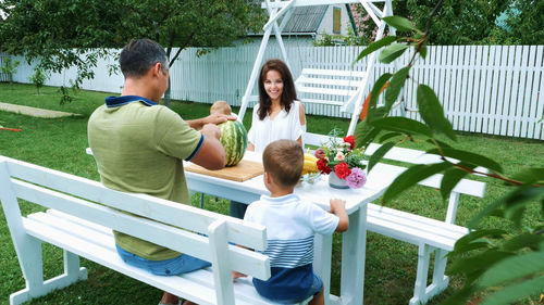 A young family with two little funny children sitting at a dinner table in the garden, in summer