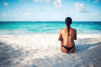 Rear view of shirtless woman sitting on sand beach against sky