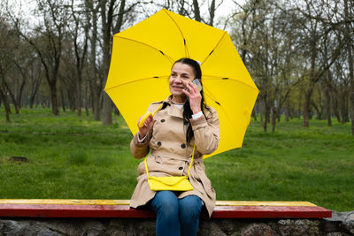 Happy senior woman in yellow rain coat with yellow umbrella is talking on a smartphone and walking 