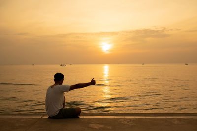 Rear view of man sitting on sea against sky during sunset