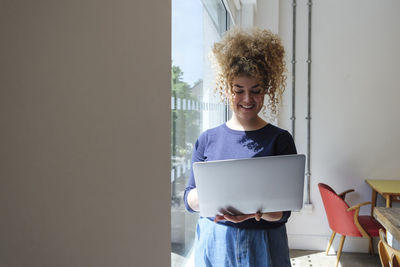 Young businesswoman standing by window holding laptop