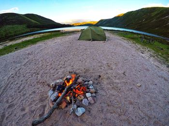 Fish-eye lens view of campfire at lakeshore against mountains