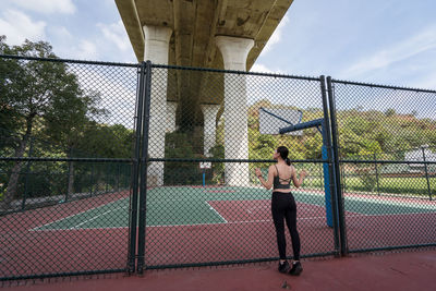 Back view of slim female athlete in sportswear standing near metal net of basketball playground in taichung