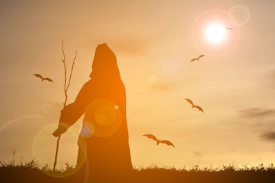 Silhouette person in halloween costume standing on field with birds flying in background during sunset