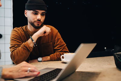 Young male hacker looking at female programmer using laptop at desk in office