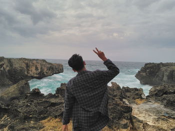 Rear view of man standing on rock by sea against sky