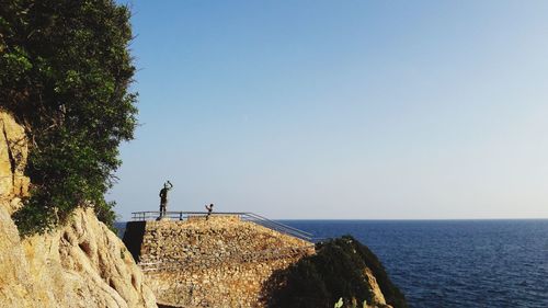 People on cliff by sea against clear sky