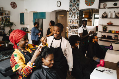 Smiling female hairdresser talking with male coworker while making locs of customer in barber shop