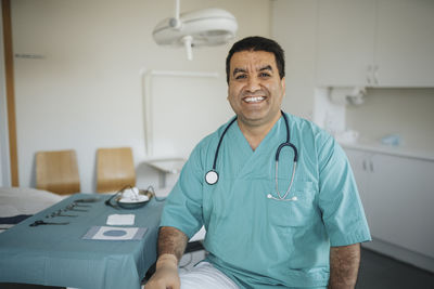 Portrait of smiling male doctor wearing scrubs sitting in medical examination room
