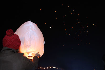 Rear view of woman holding illuminated paper lantern against sky at night