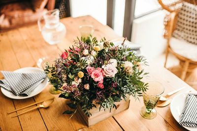 From above bouquet of miscellaneous flowers and green plant twigs in a wooden box on a timber table set for a meal with beautiful designed rattan chair on the background