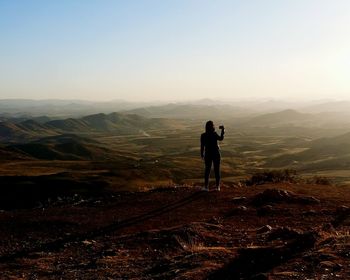 Rear view of silhouette mid adult woman photographing with mobile phone while standing on mountain against clear sky