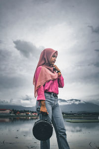 Teenage girl with bag wearing hijab standing by lake against sky