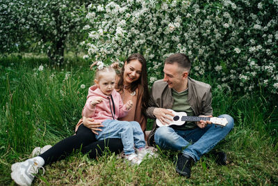 Family dad mom baby daughter in the garden blooming apple trees, father playing the ukulele