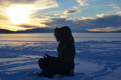Woman sitting on snow covered beach against sky during sunset
