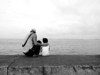 Rear view of father and son sitting on retaining wall against sky