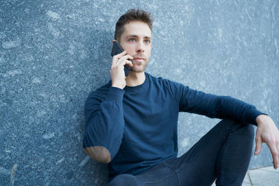 Side view of modern male in casual wear sitting near stone wall and br