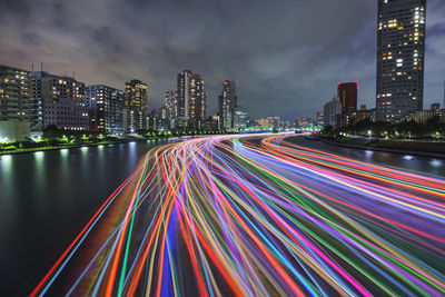 Light trails on city by buildings against sky at night