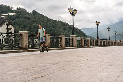Young man in protective equipment riding on roller skates along embankment against  forest mountains