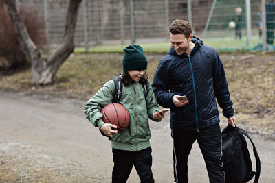 Happy father and son using mobile phones while walking on street after basketball practice in winter