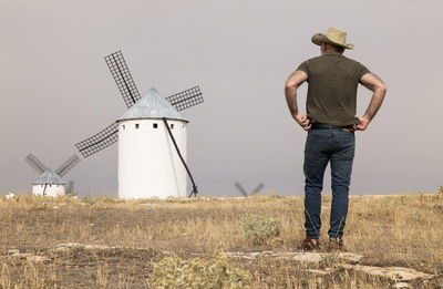 Rear view of man with cowboy hat standing on field with spanish white windmills against sky
