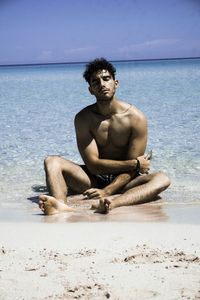 Full length of shirtless man sitting on shore at beach during sunny day