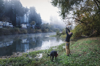 A man is holding a baby near a river and a dog