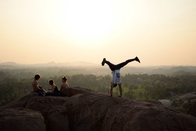 Man doing handstand by friends on rock against sky