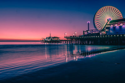 Illuminated ferris wheel by sea against sky at sunset