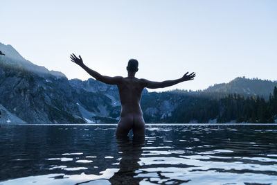 Rear view of naked man standing with arms outstretched in lake against clear sky