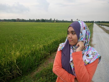 Woman in hijab looking away while standing on country road by rice paddy field