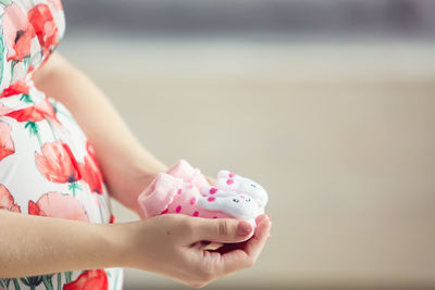 Pregnant woman holding a set of pink newborn shoes on a blurred beige background