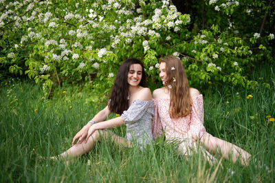 Two young girls in dresses are sitting on the green grass under a white tree and laughing