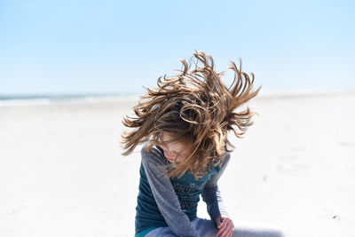 Happy girl shaking head while sitting at beach against sky