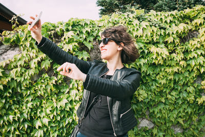Smiling woman taking selfie with smart phone while standing against plants at park